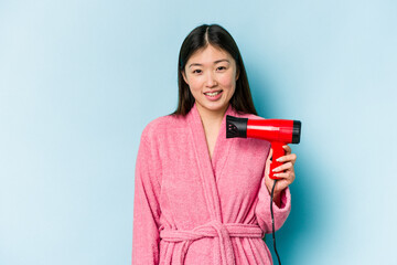Young asian woman wearing a bathrobe and holding hairdryer isolated on pink background laughing and having fun.