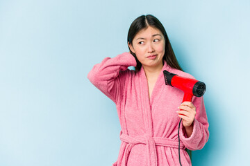 Young asian woman wearing a bathrobe and holding hairdryer isolated on pink background touching back of head, thinking and making a choice.