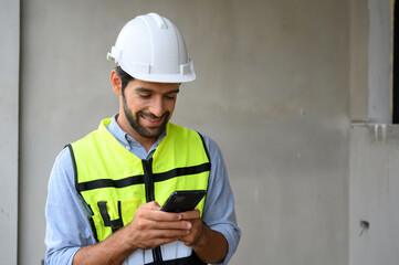 portrait of young engineer in vest with white helmet standing on construction site, smiling and holding smartphone for worker, internet, social media.