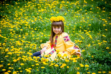 little beautiful girl  in ukrainian yellow embroidery and wreath of flowers sitting on green and...