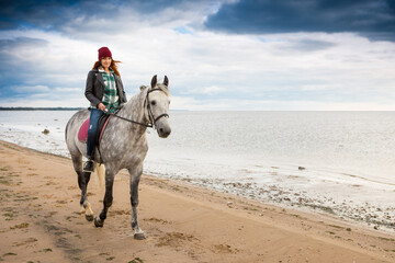 wearing jeans and jacket woman rides a gray horse along coast