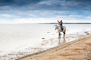 wearing jeans, warm hat and jacket female rider rides on horseback along sea shore