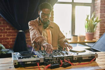 Black man in stylish casualwear turning mixers of music equipment while standing by table in loft...