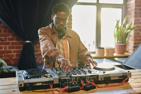 Black man in stylish casualwear turning mixers of music equipment while standing by table in loft apartment and touching turntable