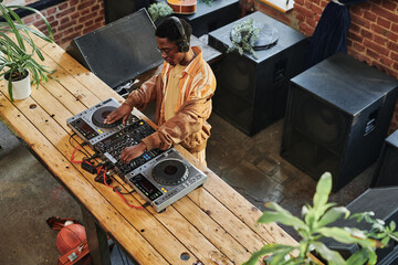 Overview of young African American male deejay standing by long wooden table with dj set and...