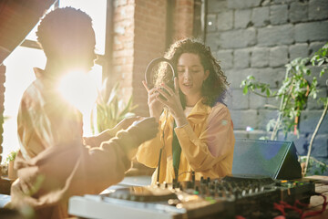 Young female in casualwear holding headphones while creating new music with her African American...