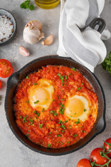Shakshuka in a frying pan on a gray concrete background with ingredients. Eggs cooked in tomato sauce with spices and fresh parsley on top. Copy space.