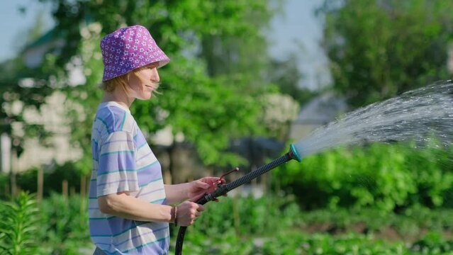 Happy female farmer watering her garden with hose in sunny summer day. Positive adult woman in funny hat pouring green plants in backyard.