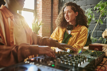 Pretty girl with dark long wavy hair talking to her African American boyfriend mixing sounds on dj...
