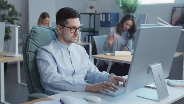 Attractive young man with glasses working on computers in the office. In the background businesspeople work. Slow-motion
