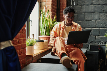 Young black man in casualwear sitting by windowsill with green domestic plants and neworking on...