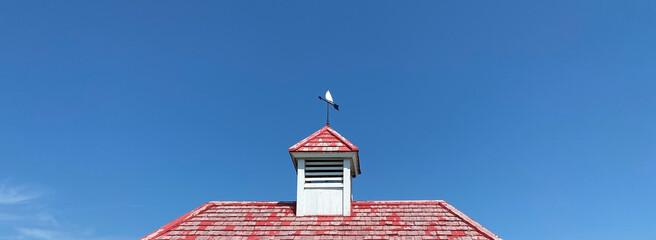 Toit en bois rouge avec une tour de clocher et une girouette pour indiquer l'orientation du vent. Ciel bleu. - obrazy, fototapety, plakaty