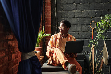 Young serious African American man with laptop sitting by windowsill and looking through window...