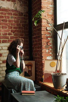 Young female with photocamera taking photo while sitting in front of window against brick wall of modern studio or loft apartment
