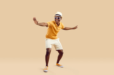 Fototapeta na wymiar Cheerful stylish african american man having fun dancing on light beige background. Funny smiling dark skinned man in sneakers, shorts, polo shirt and hat with sunglasses. Full length. Banner.