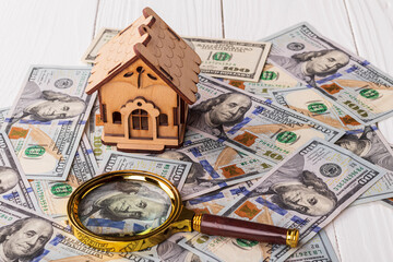 A toy house made of wood with a roof costs money, on a hundred-dollar bill, next to a magnifying glass. Concept: mortgage, purchase, sale of real estate, rental housing