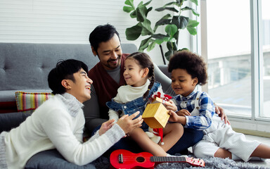 Couple male gay giving birthday gift, taking care with love to adopted children who are little diverse Caucasian and African girl and boy, sitting on sofa in living room at home. LGBT, kids Concept.