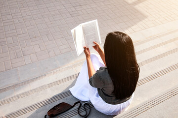 Young woman seating on stairs and reading her favourite book in a nice sunny summer day. Concept of...