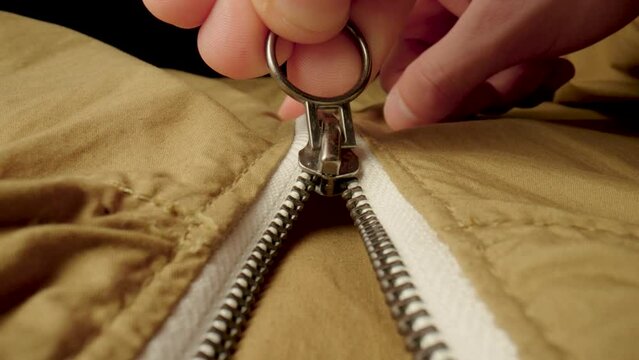 A man fastens a metal zipper on a beige jacket in macro. Extreme close up zipper with hand fastens clothing, while camera following the man hand. Fashion, textile and clothing industry concept.