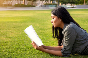 Fototapeta premium Close up portrait of young Indian ethnicity beautiful girl reading a book, laying on a grass