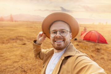 Happy hiker man with hat making selfie photo portrait background mountain Altai sunset. Concept travel blog, adventure life trip