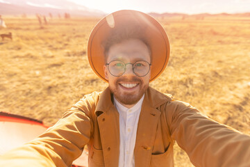 Happy blogger man with hat making selfie photo portrait background red tent and mountains. Concept hiking travel adventure