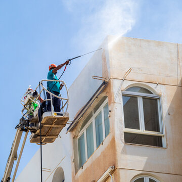 Hombre en una gr&uacute;a realizando trabajos de limpieza de fachada con agua a presi&oacute;n en un edificio alto.