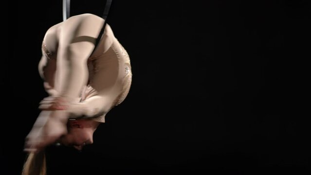 Flexible young Caucasian woman hanging on air hoop holding feet with hands spinning. Skilled slim aerial acrobat performing modern dance at black background. Art and beauty concept
