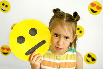 Sad upset girl pouting looks at the camera holding a carton saddened emoticon. Various emojis on a white background - obrazy, fototapety, plakaty