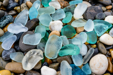 Natural polish textured sea glass and stones on the seashore. Azure clear sea water with waves. Green, blue shiny glass with multi-colored sea pebbles close-up. Beach summer background