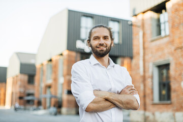 Portrait of young handsome smiling bearded man with crossed arms on background of city. Charming Caucasian businessman standing near office space during break.