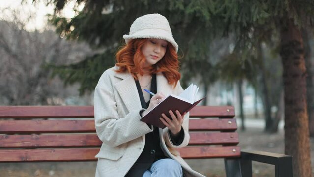 A young Asian-looking poet with red hair is sitting on a bench in a city park and composing poetry. She writes down all her options in a notebook. A woman dressed in a light coat and hat. Slow motion.