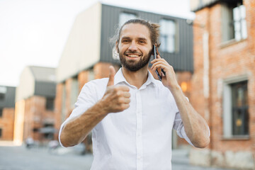 Portrait of smiling young man doing a phone call showing thumb up. Handsome sporty bearded dark-haired man is using the smart phone outdoor.