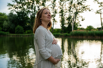 Beautiful pregnant young woman outside, relaxing and enjoying life in nature, warm sunny picture. Lovely couple near the pond. Maternity photoshoot. Young man standing with his pregnant woman