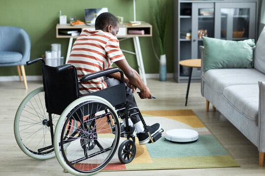 Full length portrait of modern black man with disability using robot vacuum cleaner and smart home technology, copy space