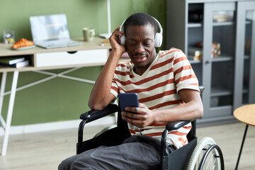 Portrait of smiling black man with disability listening to music and using phone in modern home, copy space