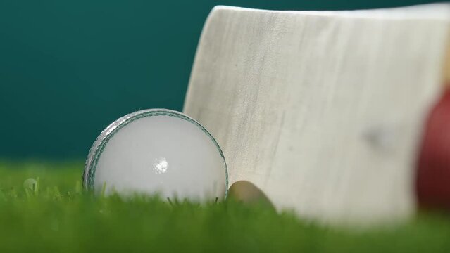 A low angle shot view zooming in to a white and green cricket ball and a wooden cricket bat with a red grip sitting in the grass.