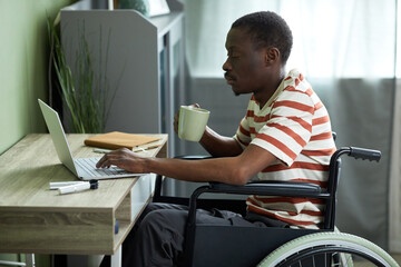 Side view portrait of African American man in wheelchair using laptop while working from home and drinking coffee