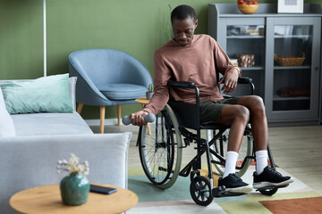 Full length portrait of young black man with disability exercising at home using dumbbell, copy...