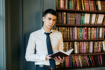 A young businessman man in a white shirt and tie is standing near bookcase, holding open book in his hands and confidently looking ahead at the camera