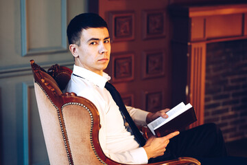A young male manager in a white shirt is sitting sideways on vintage armchair infront of fireplace, holding open book in hands and confidently looking at the camera with his head turned to the right