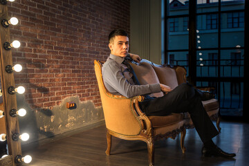 A young businessman in trousers, shirt and tie is sitting on old vintage sofa with his hand on the armrest, against the background of brick wall and thoughtfully looks at camera