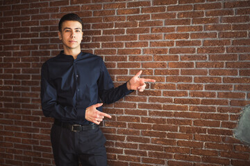 A young businessman in trousers and blue shirt is standing against brick wall, pointing his fingers to the right side and looking forward