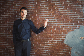 A young businessman in trousers and a blue shirt stands against a brick wall, stretched out his left hand to the side and looks forward
