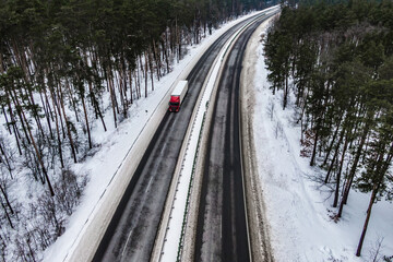 Fototapeta premium red truck on the higthway. cargo driving by road seen from the air. Top view landscape. shooting from a drone. Cargo delivery in winter. Aerial view of snow covered road in winter forest