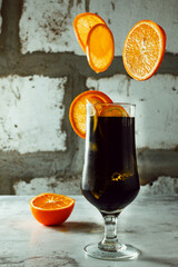A tall glass of a dark drink, with flying circles of juicy orange, levitating fruits, a cocktail on a background of concrete and cement, bright orange