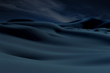 Desert hills and dunes at night under the stars and cloudy sky in blue atmosphere.
