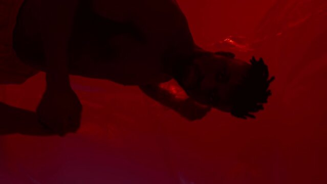 Young very active and energetic african american man smiling and dancing in good mood on dark background with red light. Vertical video
