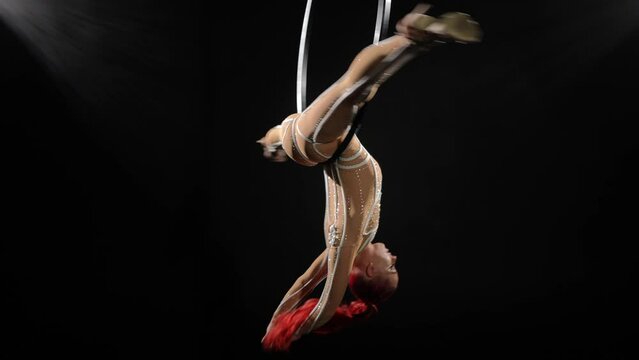 Wide shot slim young woman in split doing gazelle hanging on air hoop at black background. Portrait of confident flexible beautiful redhead Caucasian performer dancing acrobatic dance on stage