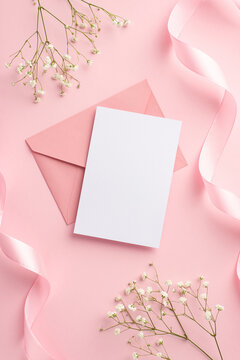 Wedding invitation concept. Top view vertical photo of pink envelope paper sheet silk curly ribbons and white gypsophila flowers on isolated pastel pink background with blank space